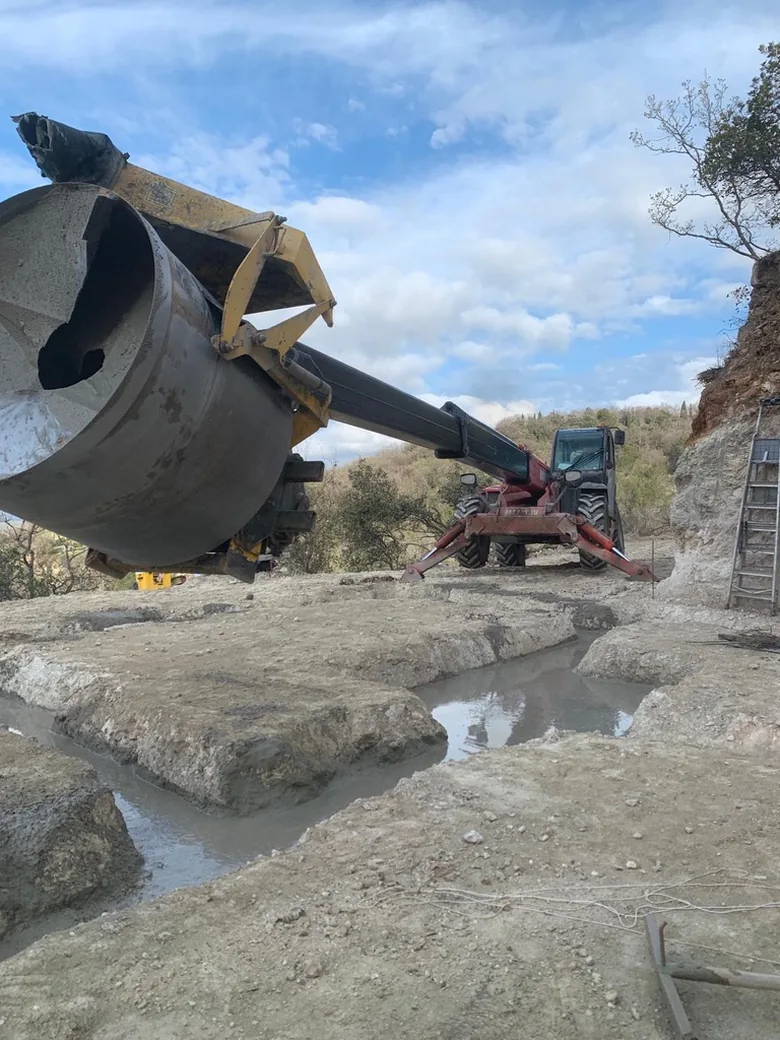 Benne à béton au-dessus d'une fouille creusée dans la roche, engin de terrassement à côté.